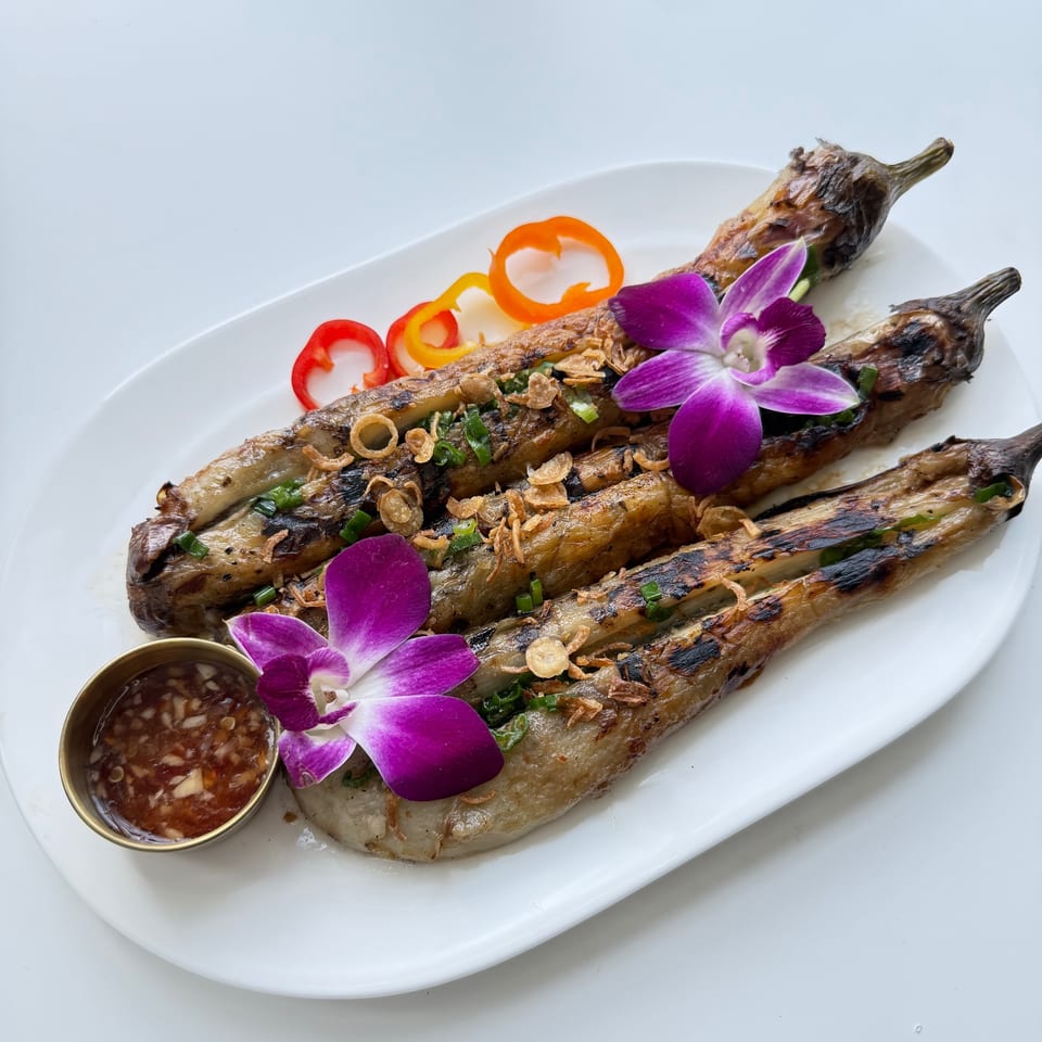 Grilled eggplants garnished with fried shallots, green onions, bell peppers, and purple orchids, with a side sauce.