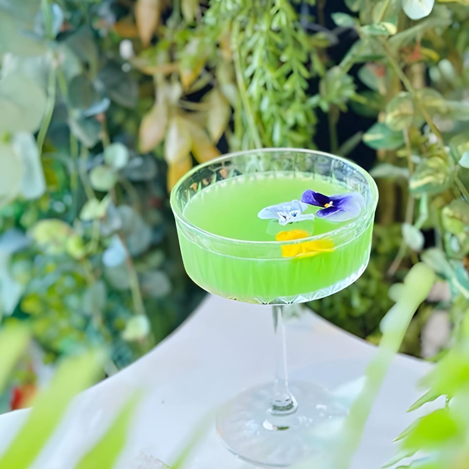 Vibrant green cocktail in a coupe glass, garnished with purple and yellow edible flowers, surrounded by lush greenery.