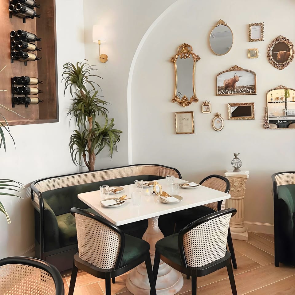 A dining area with a white table, dark green booth, cane chairs, and gold-framed mirrors on the wall.