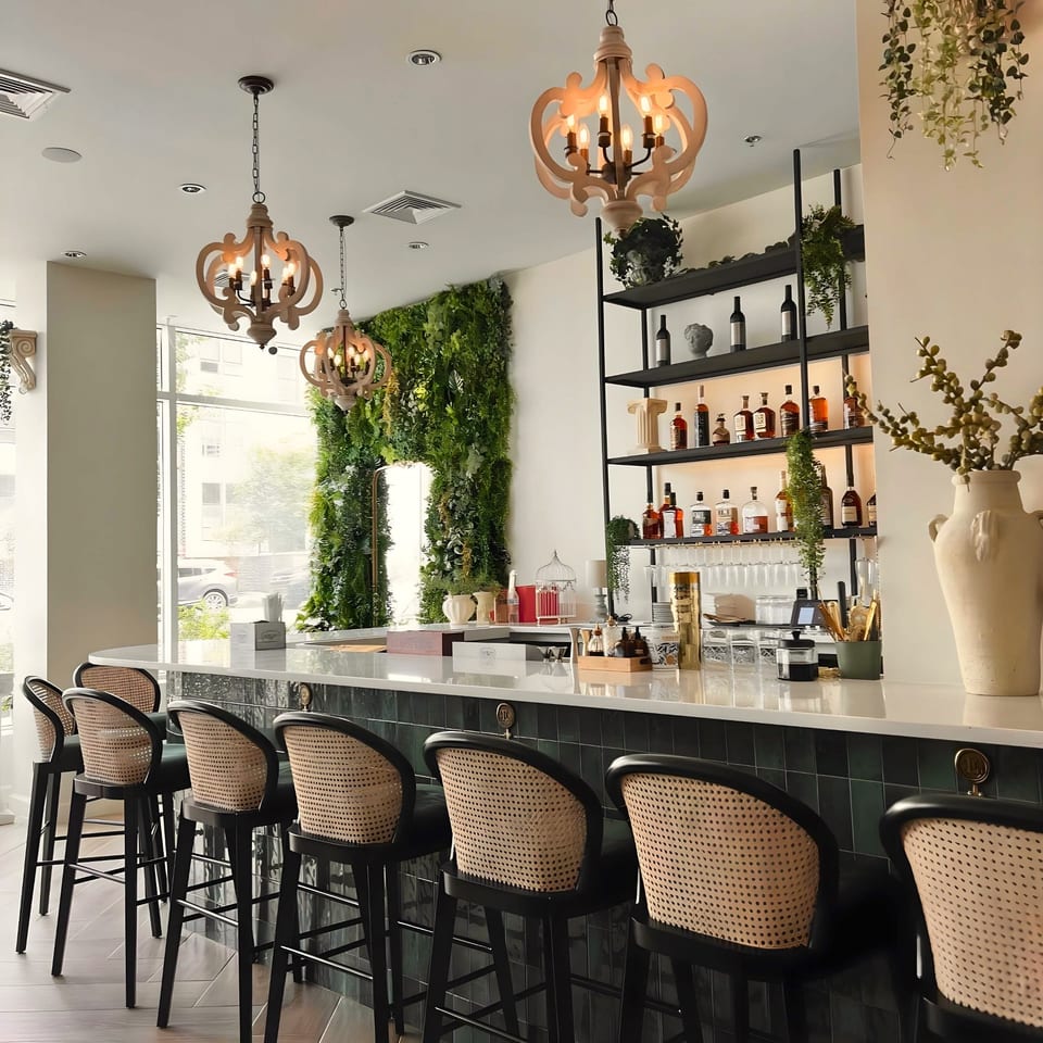 Lantern House Kitchen & Bar's elegant bar with rattan stools, a green wall, and decorative chandeliers.
