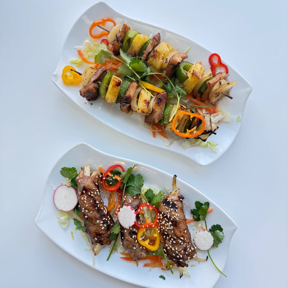 Two plates of Asian-fusion skewers: one with grilled meat and pineapple, the other with sesame-crusted beef ribs.